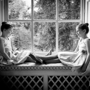 Black and white window-lit image of young flower girls chatting in window at Hartwell House Hotel wedding