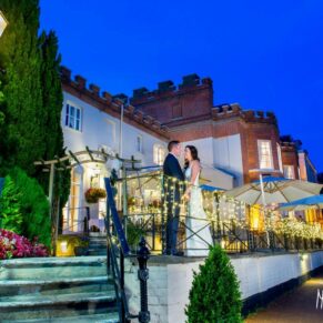 Bride and groom on terrace at Taplow House Hotel at dusk with fairy lights and pink flowers in foreground