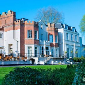 Taplow House Hotel on a sunny summer day, with tables, chairs and umbrellas laid out on the terrace and green grass and shrubs in foreground