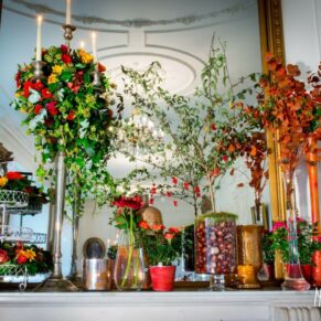 Autumnal wedding breakfast place setting with chestnuts, yellow and red berries, and orange and red roses in jars and ivy with bronze-sprayed eucalyptus branches