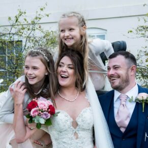Photography of the wedding bridesmaids giving a big hug to the bride and groom at the front of Sopwell House Hotel