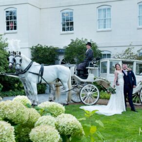 Photography of the newlyweds posing for a picture at the front of their Sopwell House wedding venue with their fabulous white horses and carriage