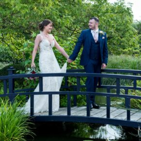 The relaxed bride and groom walk over the bridge hand in hand in the lush green gardens of Sopwell House