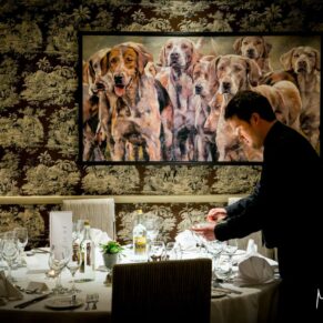 Photography of a member of staff setting up a table in the dining area whilst stood under a huge painting at the Bay Tree Hotel in Burford