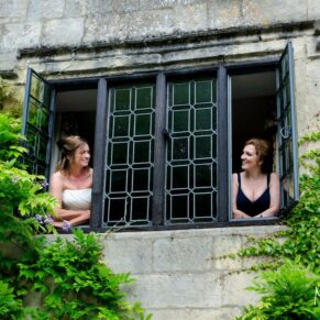 Photography of the bride and her bridesmaid giggling in the window whilst surrounded by lush wisteria at The Bay Tree Hotel in Burford on her wedding day