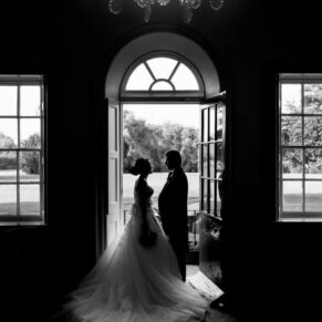 Silhouette pose of the newlyweds in a feature doorway with a huge twinkling chandelier above in one of the Bisham Abbey's fabulous rooms
