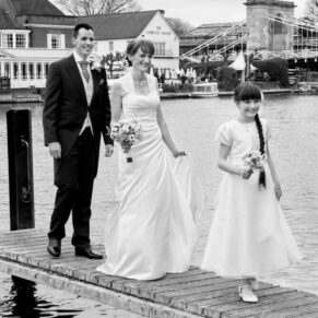 The newlyweds with their flower girl walking on the jetty with their hotel seen across the water