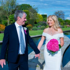 The bride and groom walking along hand in hand through the lush green gardens of The Grove in Watford