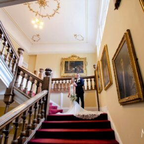 The bride and groom pose on the grand staircase surrounded by stunning historic paintings at their Farnham Castle wedding