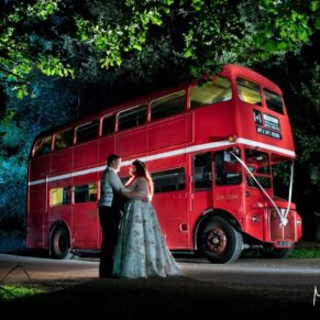The bride and groom pose beside their red double decker London bus at dusk at their evening reception  