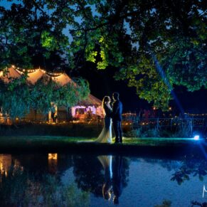 A floodlit silhouette pose of the newlyweds captured over the lake with their marquee in the background