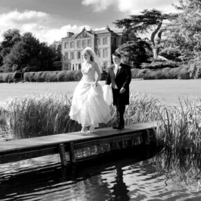 The happy newlyweds cross the narrow wooden bridge at their wedding with fabulous views of the grounds and main house behind them