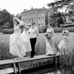 The bride and bridesmaids make their way across the narrow wooden bridge as they arrive for the outdoor ceremony with distant views of the main house behind