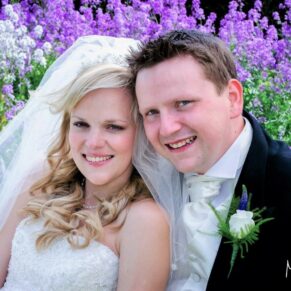 The happy newlyweds in a close-up pose looking away from the camera with mauve flowers behind them