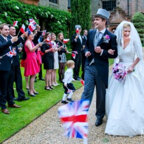 The smiling bride and groom are greeted by their enthusiastic flag waving guests as they arrive from their church ceremony at their Chenies Manor wedding