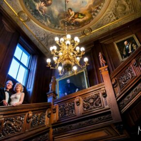 The bride and groom pose on the dramatic grand staircase at Cliveden House under its painted ceiling and huge chandelier