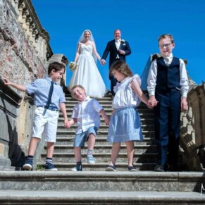 The couple walk down the steps with their children on the South Terrace of Cliveden House on their glorious blue sky wedding day