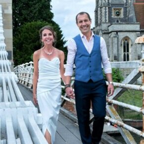 The bride and groom walking across Marlow Bridge with All Saints Church behind them as they approach their drinks reception