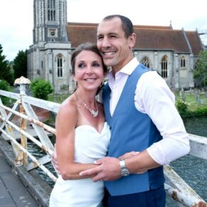 The smiling newlyweds on the bridge with All Saints Church behind them for their Compleat Angler wedding in Marlow