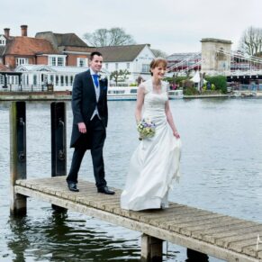 The happy bride and groom walking back along the jetty after their wedding ceremony with Marlow Bridge and The Compleat Angler off in the distance over the water