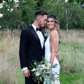 The bride and groom pose for the camera under their flower archway in the meadow surroundings