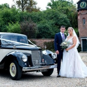The newlyweds pose beside their car under the clock tower at the Tudor Barn
