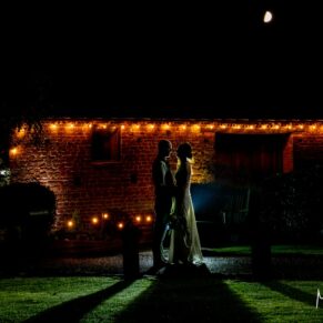 The newlyweds pose for the camera at night-time outside the illuminated barn at their Dovecote Events wedding