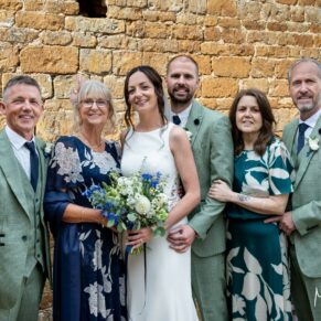 The newlyweds with their parents pose for the camera next to a rustic wall