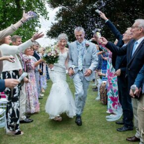 Photography of the smiling newlyweds run through the colourful confetti aisle after their Bisham Abbey outdoor wedding civil ceremony next to the River Thames