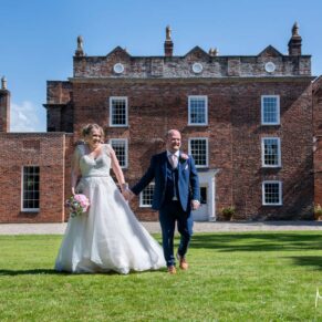 The bride and groom walk along in front of Meols Hall on their gloriously sunny wedding day